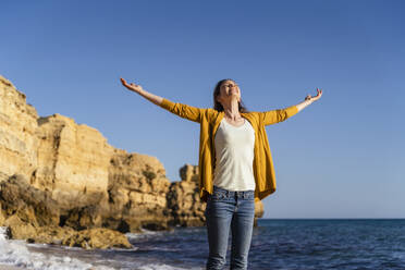 Smiling woman with arms outstretched standing at beach on sunny day - DIGF17860