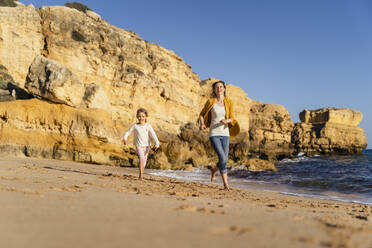 Smiling mother and daughter running on shore at beach - DIGF17863