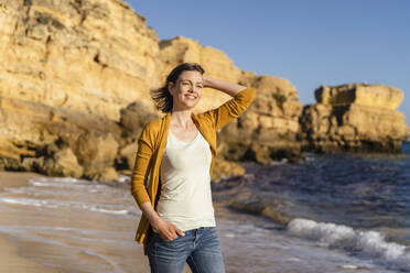 Smiling woman with hand in hair standing at beach on sunny day - DIGF17875