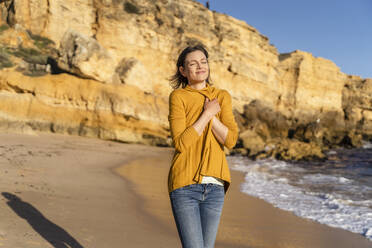 Smiling woman wearing shrug standing in front of rock formation on shore at beach - DIGF17876