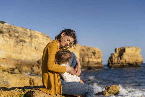 Smiling woman with daughter sitting on rock at beach - DIGF17880