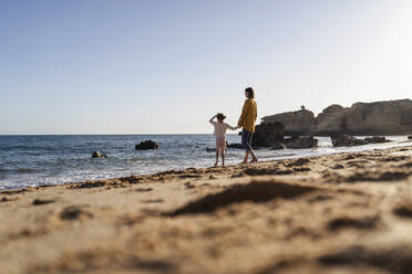 Mother and daughter holding hands standing on shore at beach - DIGF17883