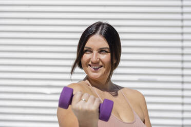 Happy curvy woman exercising with dumbbell exercising in front of corrugated wall - EIF04069
