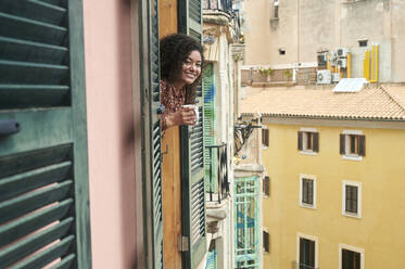 Smiling young woman holding coffee cup leaning out of window - KIJF04442