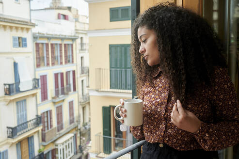 Young woman holding coffee cup looking through window at home - KIJF04443