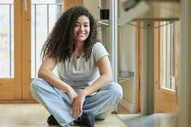 Lächelnde junge Frau sitzt im Schneidersitz auf dem Boden zu Hause - KIJF04445
