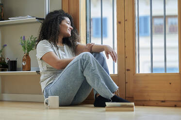 Smiling woman looking through glass door sitting on floor at home - KIJF04448