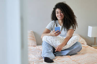 Smiling woman holding mobile phone sitting on bed at home - KIJF04450