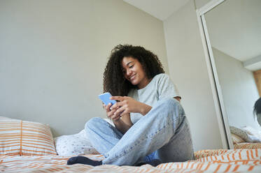 Smiling woman using smart phone sitting on bed at home - KIJF04451