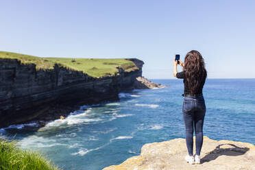 Junge Frau, die mit ihrem Handy auf einem Felsen stehend das Meer fotografiert - EGHF00382