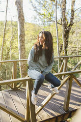 Lächelnde junge Frau sitzt auf dem Holzgeländer einer Brücke - EGHF00385