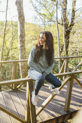 Smiling young woman sitting on wooden railing of bridge - EGHF00385