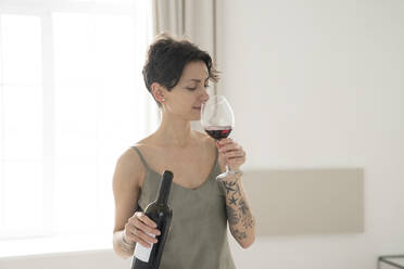 Frau hält Flasche riechendes Glas Wein stehend zu Hause - VPIF05966