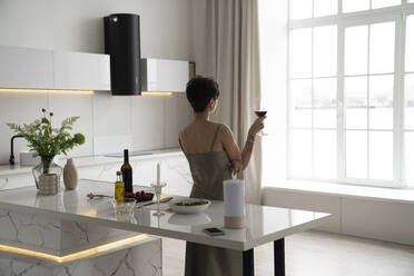 Frau hält Weinglas und schaut durch das Fenster in der Küche zu Hause - VPIF05968