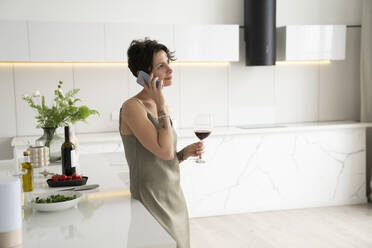 Frau, die ein Weinglas hält und mit einem Mobiltelefon spricht, steht in der Küche - VPIF05969