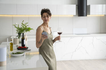 Woman holding wineglass using smart phone standing in kitchen - VPIF05970