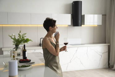 Happy woman holding wineglass talking through mobile phone speaker in kitchen - VPIF05971