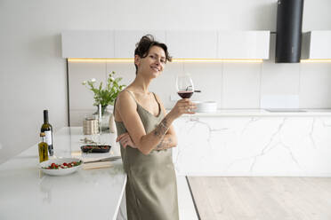 Smiling woman holding wineglass leaning on kitchen island at home - VPIF05978