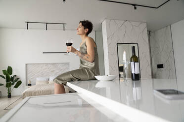Lächelnde Frau, die ein Weinglas hält, sitzt auf einer Kücheninsel zu Hause - VPIF05989