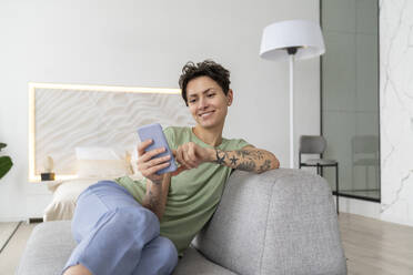 Smiling woman using mobile phone sitting on sofa at home - VPIF05998