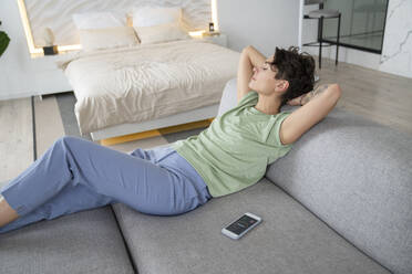 Woman with hands behind head resting on sofa at home - VPIF05999