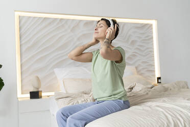 Frau mit geschlossenen Augen, die zu Hause auf dem Bett sitzend Musik über drahtlose Kopfhörer hört - VPIF06011