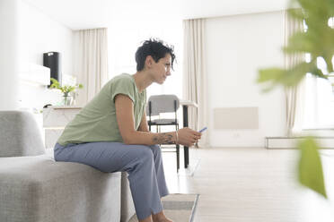 Woman holding smart phone sitting on sofa at home - VPIF06014