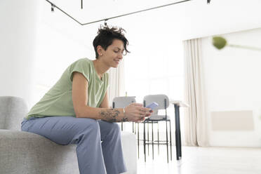 Smiling woman using smart phone sitting on sofa at home - VPIF06015