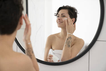 Happy woman applying moisturizer looking at mirror in bathroom - VPIF06023
