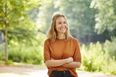 Smiling blond woman with arms crossed standing in park - JHAF00151