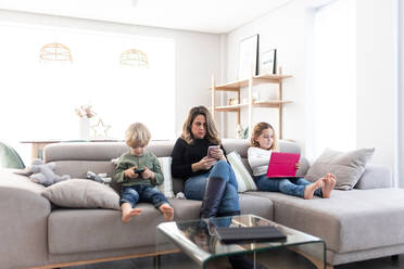 Focused mother with children browsing smartphones and tablet while sitting together on comfortable sofa in light living room at home - ADSF34568