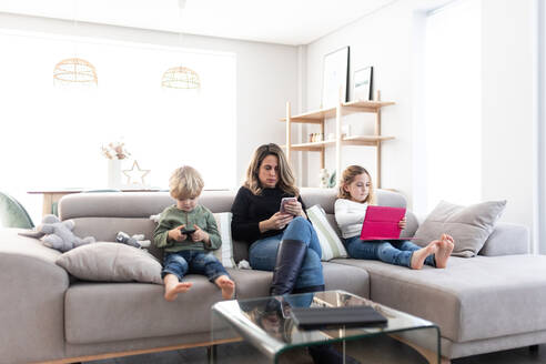 Konzentrierte Mutter mit Kindern, die mit Smartphone und Tablet surfen, während sie zusammen auf dem bequemen Sofa im hellen Wohnzimmer sitzen - ADSF34568