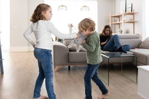 Wütende Schwester versucht, dem Bruder das Spielzeug wegzunehmen, während sie im hellen Wohnzimmer in der Nähe der Mutter auf der Couch streitet - ADSF34569