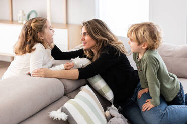 Caring mother and daughter looking at each other while sitting on comfortable couch with cute son in light living room - ADSF34573