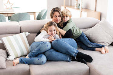 Full body of caring mother resting on comfortable sofa with positive children while spending time together in light living room - ADSF34574