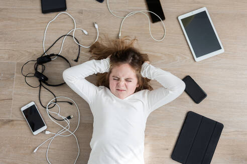 Draufsicht auf ein unglückliches Mädchen, das auf dem Boden in der Nähe einer Fülle von modernen digitalen Geräten und USB-Kabeln in einem hellen Raum liegt und in die Kamera schaut - ADSF34575