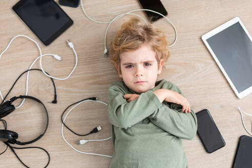 Draufsicht auf einen unglücklichen Jungen, der auf dem Boden in der Nähe einer Fülle von modernen digitalen Geräten und USB-Kabeln in einem hellen Raum liegt und in die Kamera schaut - ADSF34578