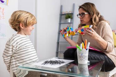 Professionelle weibliche Psychologin zeigt einem Jungen, der am Tisch sitzt, bunte Spielsachen mit gezeichneten schwarzen Strichen während eines Termins in einem hellen Raum - ADSF34580