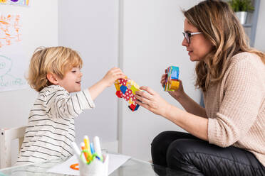 Side view of female psychologist showing colorful toys to cute boy while sitting at table in light room during appointment - ADSF34583