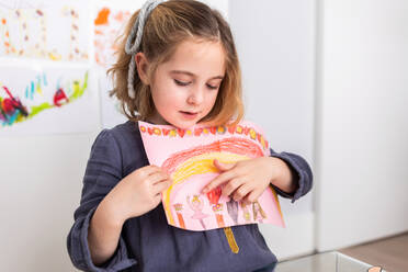 Cute girl pointing at colorful drawing sitting at table in light room - ADSF34588