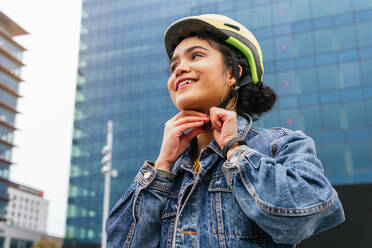 Von unten lächelnd junge kolumbianische Frau in stilvollem Denim-Outfit Befestigung Schutzhelm vor dem Radfahren auf der Straße der modernen Stadt - ADSF34593