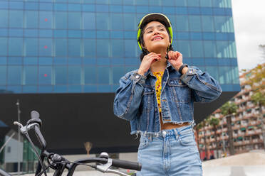 From below smiling young Colombian female in stylish denim outfit fastening protective helmet before riding bike on street of modern city - ADSF34594