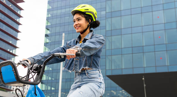 Von unten Seitenansicht der fröhlichen kolumbianischen jungen Dame trägt trendige Denim-Kleidung und Schutzhelm Reiten ein Fahrrad in der Nähe von modernen Glasgebäude während der aktiven Hobby Praxis - ADSF34596