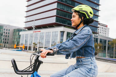 Fröhliche kolumbianische junge Frau in trendiger Jeans und mit Schutzhelm auf dem Fahrrad in der Nähe eines modernen Glasgebäudes bei der Ausübung eines aktiven Hobbys - ADSF34597