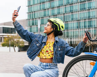 Lächelnde kolumbianische Frau in Jeanskleidung und Schutzhelm, die ein Selfie mit ihrem Smartphone macht, während sie eine Pause beim Fahrradfahren auf der Straße macht - ADSF34598