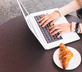 High angle of crop anonymous female remote worker using netbook for online project while working on street cafe at table with croissant - ADSF34602