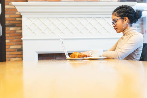 Seitenansicht eines weiblichen Fernarbeiters, der ein Netbook für ein Online-Projekt verwendet, während er in einem Straßencafé an einem Tisch mit Croissant arbeitet - ADSF34603