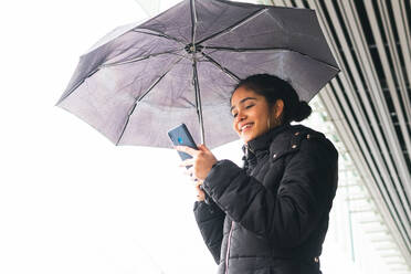 Von unten fröhliche Kolumbianerin in Oberbekleidung, die mit Regenschirm auf der Straße steht und in ihrem Handy blättert - ADSF34606