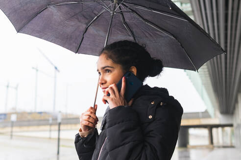 Fröhliche Kolumbianerin in Oberbekleidung mit Regenschirm beim Telefonieren mit dem Handy auf der Straße - ADSF34607