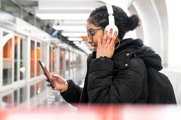 Side view of Colombian female in outerwear and eyeglasses scrolling social media on cellphone while listening to music in wireless headphones and waiting train on subway station - ADSF34611